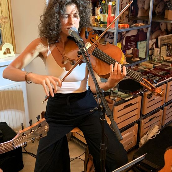 Eine Frau spielt die Geige, während sie in einem Raum steht. Sie trägt ein weisses ärmelloses Oberteil und schwarze Hose. Hinter ihr befinden sich Regale mit verschiedenen Gegenständen.