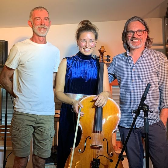 Drei Personen posieren mit einem Cello für ein Foto. Die Frau in Blau hält das Cello. Der Mann links trägt ein weißes T-Shirt und grüne Shorts. Der Mann rechts trägt ein blaues Hemd und Hose.