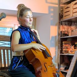 Eine Person in einem blauen Kleid sitzt auf einem Holzstuhl und spielt ein Cello mit einem Mikrofon davor.