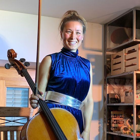Eine Frau in einem blauen Kleid hält ein Cello. Sie steht in einem Raum mit einem Holzstuhl, einem Regal mit verschiedenen Gegenständen und einem Fenster mit Jalousien.