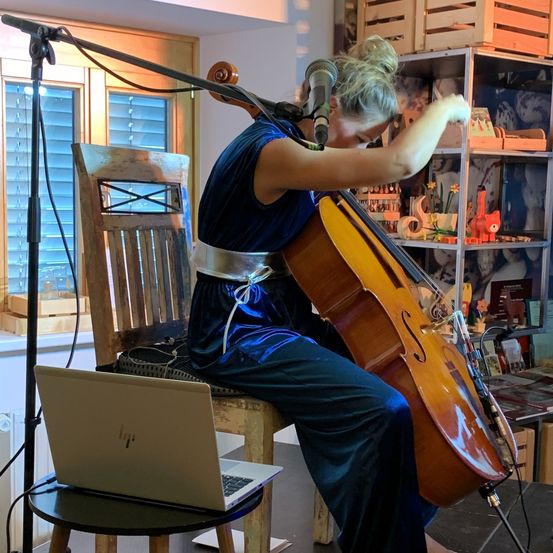 Eine Frau sitzt auf einem Stuhl und spielt Cello. Vor ihr steht ein Mikrofon und ein Laptop ist auf einem Hocker abgestellt. Der Raum hat einen Holzboden und Regale mit verschiedenen Gegenständen.