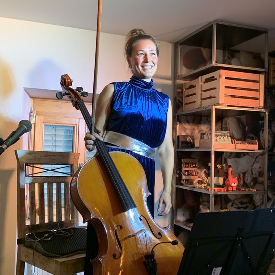 Eine Frau steht in einem Raum und hält ein Cello, mit einem Stuhl und Mikrofon in der Nähe. Hinter ihr befindet sich ein Regal mit verschiedenen Gegenständen. Sie ist in ein blaues Kleid gekleidet.