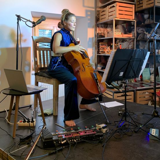 Eine Frau sitzt barfuß auf einem Stuhl und spielt Cello. Ein Notenständer mit Noten ist in der Nähe. Mikrofon und Laptop sind vor ihr aufgestellt. Im Hintergrund befinden sich Lagerboxen und ein Plüschtier.