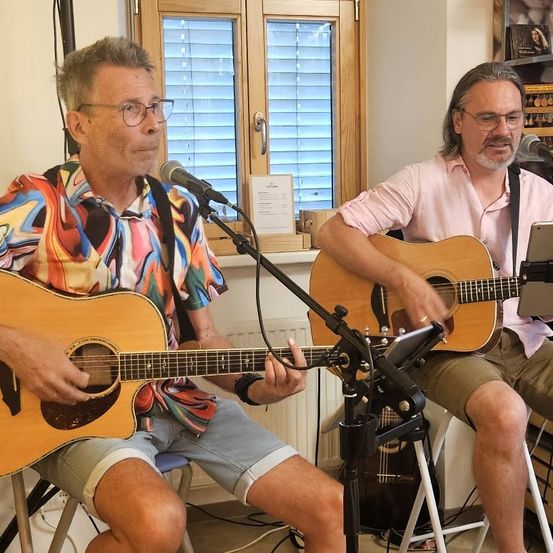 Zwei Männer sitzen auf Hockern, einer mit einem bunten Shirt, und spielen Gitarren. Sie haben Mikrofone vor sich, und einer hat ein Tablet auf dem Schoß. Sie sind in einem Raum mit einem Fenster und einem Regal mit Gegenständen.