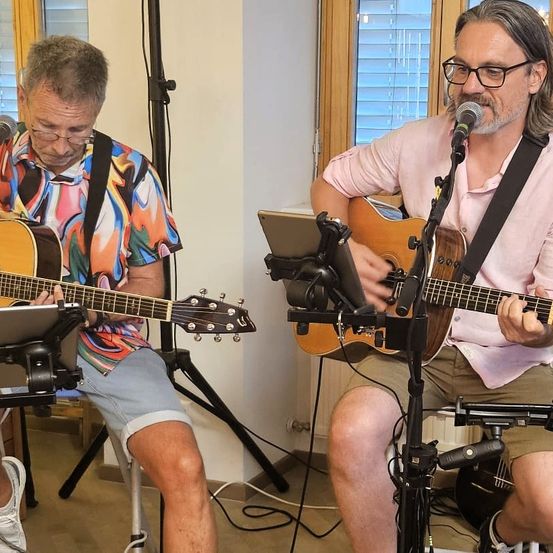 Zwei Männer sitzen und spielen Gitarren. Einer trägt ein farbenfrohes Hemd und Shorts, der andere ein rosa Hemd und eine Brille. Beide spielen in Mikrofone auf Ständern in einem Raum mit Holzboden.