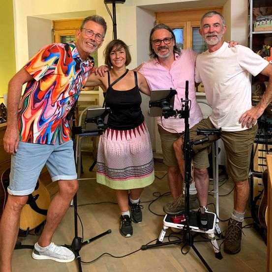 Vier Personen stehen in einem Raum mit Gitarren im Hintergrund. Eine Frau und ein Mann halten ein Tablet. Zwei Männer posieren mit ihren Armen umeinander geschlungen.