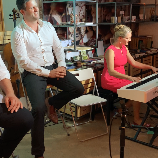 Ein Mann in einem weißen Hemd und schwarzen Hosen sitzt in einem Stuhl und lächelt. Eine Frau in einem rosa Kleid spielt Klavier in der Nähe. Ein Cello steht hinter dem Mann.