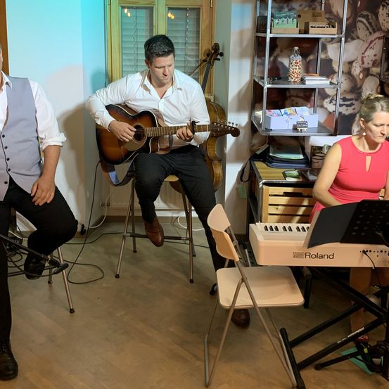 Drei Musiker spielen in einem Raum. Ein Mann spielt Gitarre, ein anderer Klavier, und der dritte steht neben ihnen. Es gibt ein Regal mit verschiedenen Gegenständen und ein Fenster mit Jalousien.