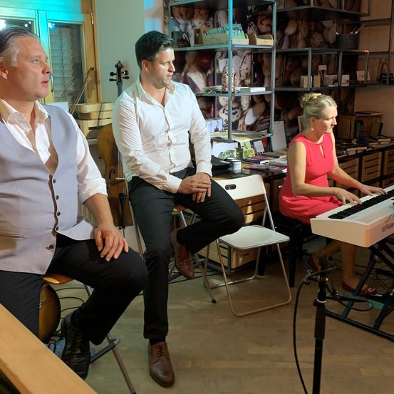 Drei Personen sind in einem Raum. Eine Frau spielt Klavier, während zwei Männer neben ihr sitzen, einer spielt Cello.