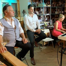 Drei Personen sitzen in einem Raum, eine spielt Klavier, eine andere mit einem Glas Wein. Zwei Männer tragen weiße Hemden und schwarze Hosen. Regale halten Spielzeug und Bücher.