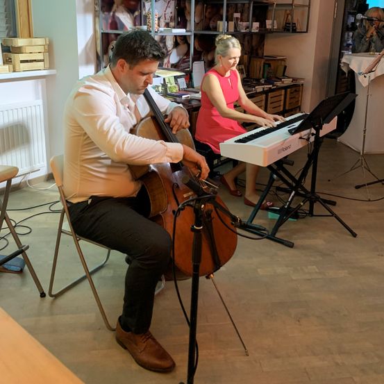 Ein Mann in einem weißen Hemd und schwarzen Hosen spielt Cello, während eine Frau in einem rosa Kleid Klavier spielt. Sie sitzen auf Stühlen in einem Raum mit Holzboden und einem weißen Heizkörper.