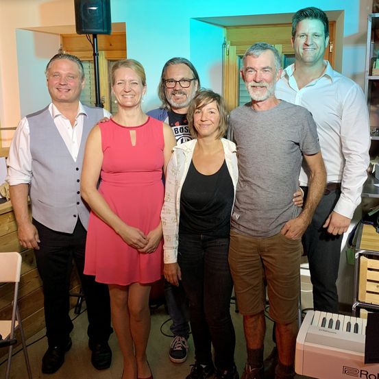 Sechs Personen, drei Männer und drei Frauen, stehen zusammen und lächeln für ein Foto. Eine Frau trägt ein rosa Kleid und eine andere Frau eine weiße Jacke. Die Männer tragen weiße Hemden, eine Weste und eine Brille. Sie befinden sich in einem Raum mit einem Klavier und einer Holzwand.