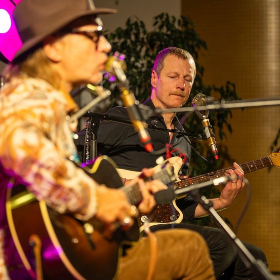 Zwei Männer spielen auf der Bühne Gitarre und singen in Mikrofone. Einer trägt Hut und Brille, der andere einen Schnurrbart. Sie stehen unter lila Licht.