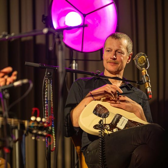 Ein Mann spielt Gitarre auf der Bühne, hält sie mit beiden Händen. Ein Mikrofon steht vor ihm, und eine pinke Lampe befindet sich dahinter.