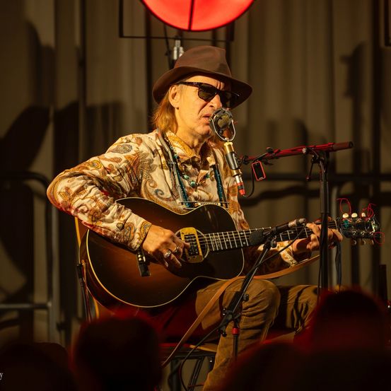 Ein Mann sitzt auf einem Stuhl, spielt Gitarre und singt in ein Mikrofon. Er trägt einen Hut, eine Sonnenbrille und ein Paisley-Hemd. Hinter ihm beleuchtet ein Scheinwerfer die Szene.