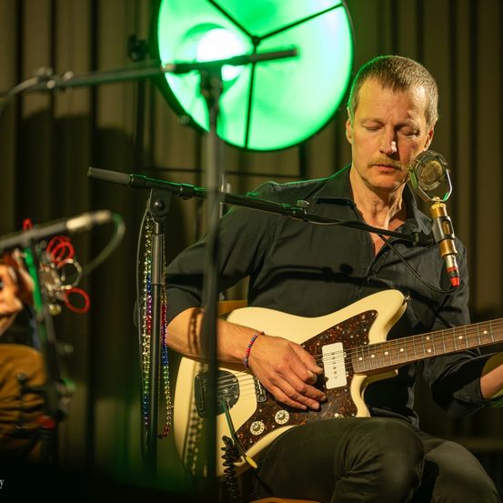 Ein Mann spielt Gitarre und singt vor einem Mikrofon auf der Bühne, wobei ein grünes Licht den Hintergrund beleuchtet.