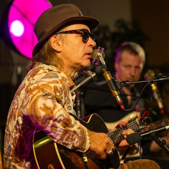 Ein älterer Mann mit Hut und Sonnenbrille singt in ein Mikrofon und hält eine Gitarre. Ein anderer Mann spielt Gitarre hinter ihm, beide unter einem rosa Licht.