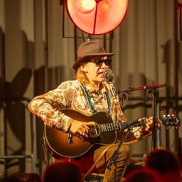 Ein älterer Mann spielt mit einer Gitarre auf der Bühne. Er trägt einen Hut und eine Sonnenbrille. Ein Mikrofon steht vor ihm. Ein Scheinwerfer ist hinter ihm.