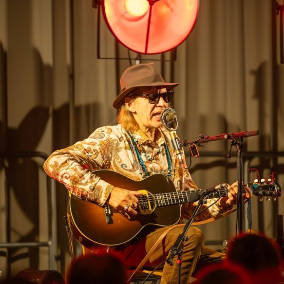 Ein älterer Mann spielt mit einer Gitarre auf der Bühne. Er trägt einen Hut und eine Sonnenbrille. Ein Mikrofon steht vor ihm. Ein Scheinwerfer ist hinter ihm.