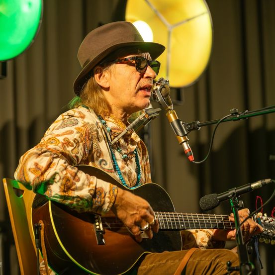 Ein älterer Mann mit Hut und Sonnenbrille spielt Gitarre und singt in ein Mikrofon auf der Bühne.