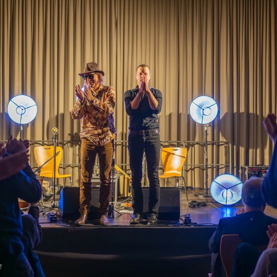 Zwei Männer stehen auf der Bühne, klatschen und lächeln. Hinter ihnen beleuchten drei blaue Lichter den Raum. Ein Publikum sitzt davor, klatscht und beobachtet.
