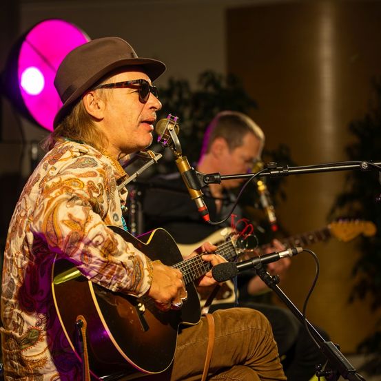 Ein Mann spielt Gitarre und singt in ein Mikrofon. Ein anderer Mann spielt Saxophon im Hintergrund. Beide sitzen vor einem Mikrofon mit Ständer. Ein rosa Licht beleuchtet den Hintergrund.