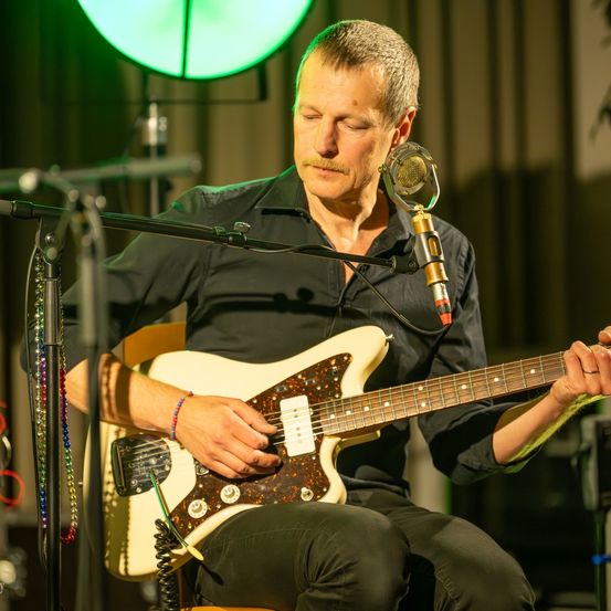 Ein Mann spielt eine weiße Gitarre mit braunem und goldenem Design, während er auf einem Hocker in einem schwach beleuchteten Raum sitzt. Er trägt ein schwarzes Hemd und eine Hose.