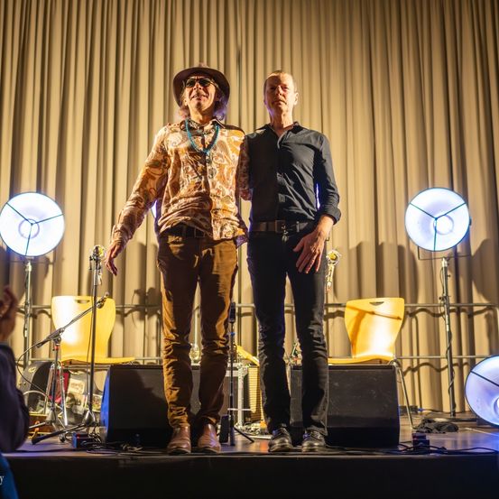 Zwei Männer auf der Bühne mit Mikrofonen, einer trägt einen Hut und eine Halskette, der andere ein schwarzes Hemd, beide sehen nach vorne, mit Musikgeräten und Lichtern im Hintergrund.