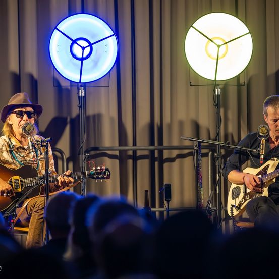 Zwei Musiker spielen auf der Bühne mit Gitarren, einer sitzt auf einem Stuhl und der andere steht, beide singen in Mikrofone mit Scheinwerfern, die sie beleuchten.