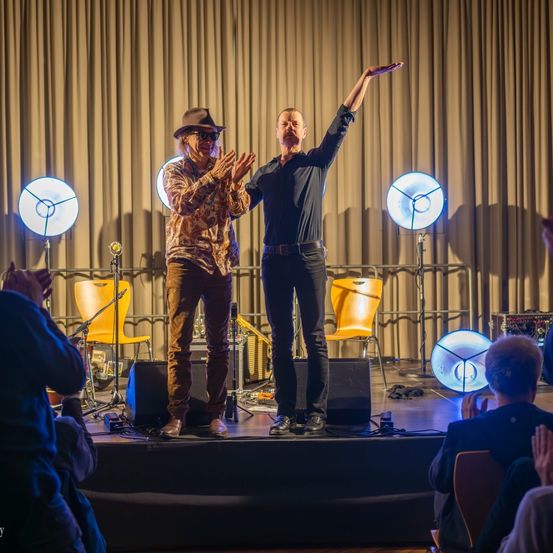 Zwei Männer stehen auf der Bühne, einer klatscht in die Hände, der andere hat seinen Arm erhoben. Sie treten vor einem Publikum auf. Es gibt Lautsprecher, Stühle und Lichter auf der Bühne. Das Publikum klatscht.