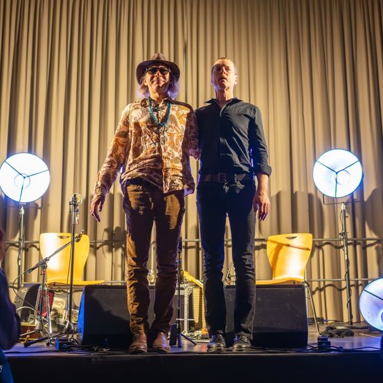 Zwei Männer stehen auf der Bühne, einer trägt einen Hut und eine Halskette, der andere ein schwarzes Hemd. Hinter ihnen stehen zwei gelbe Stühle, ein Mikrofon und Lautsprecher. Scheinwerfer beleuchten die Bühne von den Seiten.