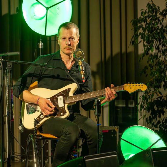 Ein Mann sitzt auf einem Stuhl und spielt eine weiße E-Gitarre unter einem grünen Licht. Er trägt ein schwarzes Hemd und eine Hose. Ein Mikrofon steht vor ihm.