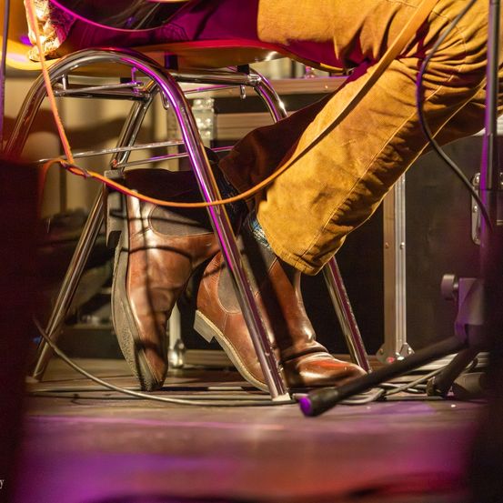 Eine Person in braunen Stiefeln und gelber Hose sitzt auf einem Hocker mit lila Beinen auf einer Bühne mit lila Beleuchtung.