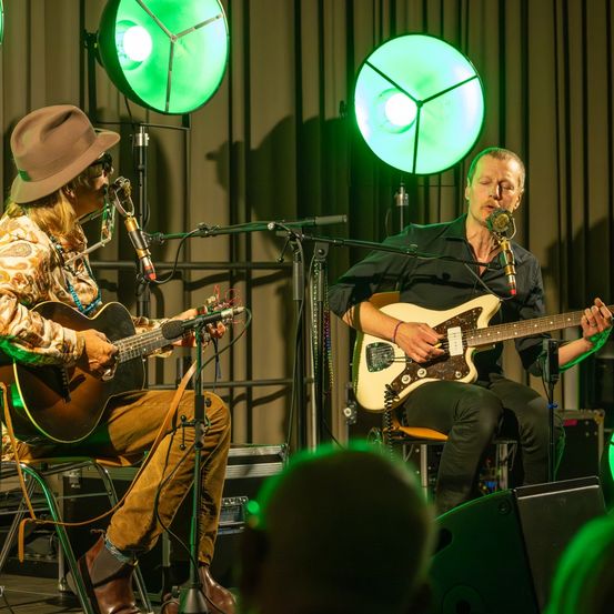 Zwei Männer spielen auf der Bühne, jeder eine Gitarre und singt in ein Mikrofon. Einer trägt einen Hut und ein Blumenhemd, der andere ein schwarzes Shirt und ein Armband. Hinter ihnen sind grüne Scheinwerfer.