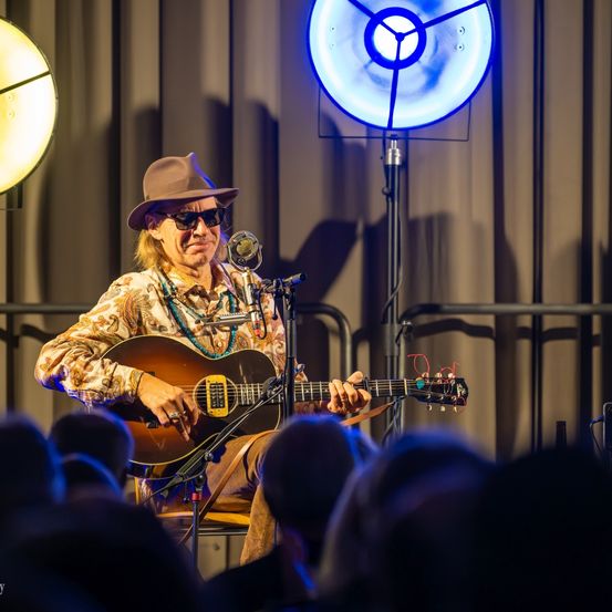 Ein Mann mit Sonnenbrille und Hut spielt mit einer Gitarre auf der Bühne, umgeben von einem großen Publikum. Zwei blaue Lichter beleuchten die Szene.