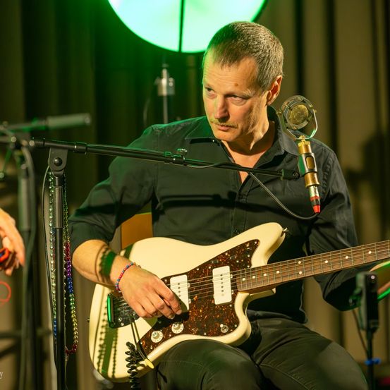 Ein Mann spielt eine Gitarre auf einer Bühne unter einem Scheinwerfer. Er sitzt vor einem Mikrofon und trägt ein schwarzes Hemd und eine Hose.