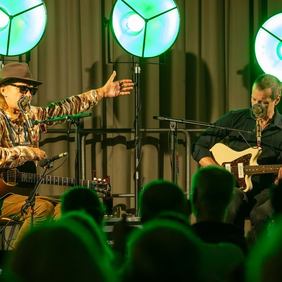 Zwei Männer spielen auf der Bühne unter grünen Lichtern. Einer spielt Gitarre, der andere singt. Das Publikum, beleuchtet von grünem Licht, schaut aufmerksam zu.