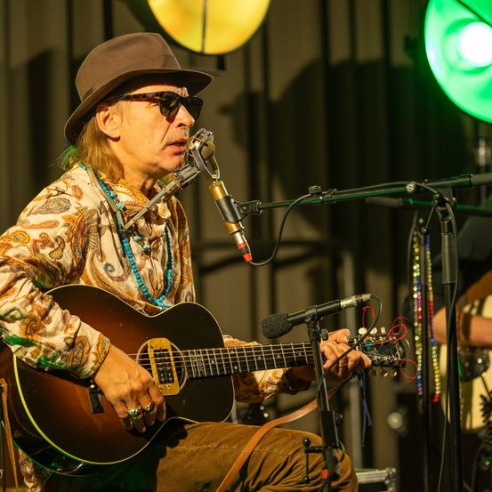 Ein Mann sitzt auf einem Stuhl, spielt eine Gitarre und singt in ein Mikrofon auf einer Bühne. Er trägt einen Hut, Sonnenbrille und mehrere Halsketten.