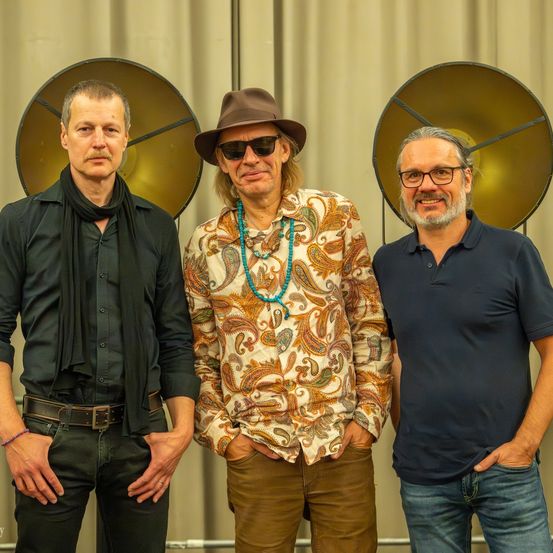 Drei Männer stehen nebeneinander, einer trägt ein Paisley-Hemd, einen Hut und eine Sonnenbrille. Ein anderer Mann trägt ein schwarzes Shirt mit einem Schal. Der dritte Mann trägt ein blaues Poloshirt und eine Brille. Hinter ihnen stehen zwei große metallische Objekte.