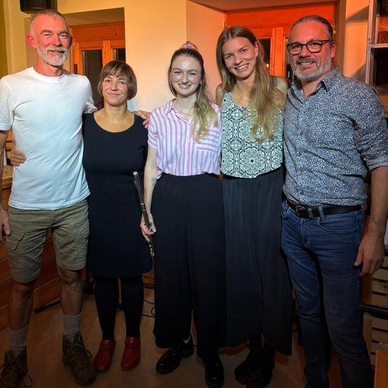 Fünf Menschen stehen für ein Foto zusammen. Zwei Frauen halten eine Flöte. Ein Mann auf der linken Seite trägt ein weißes T-Shirt und Khaki-Shorts. Ein anderer Mann auf der rechten Seite trägt eine Brille und ein gemustertes Hemd.