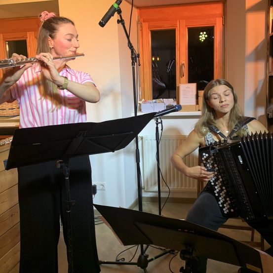 Zwei Frauen spielen Musik in einem Raum mit Holzboden. Die Frau auf der linken Seite spielt Flöte und hat einen Notenständer vor sich. Die Frau auf der rechten Seite spielt Akkordeon und hat ein Mikrofon.