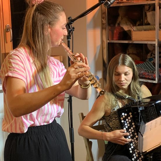 Zwei Frauen spielen gemeinsam Musik, eine spielt Flöte und die andere Akkordeon, auf einem Stuhl vor einem Mikrofon sitzend.