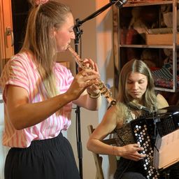 Zwei Frauen spielen gemeinsam Musik, eine spielt Flöte und die andere Akkordeon, auf einem Stuhl vor einem Mikrofon sitzend.