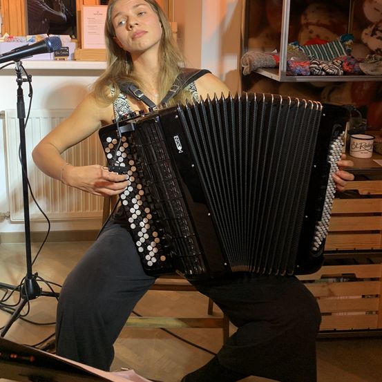 Eine Frau spielt ein Akkordeon, während sie auf einer Bank sitzt, mit einem Mikrofon vor ihr und Regalen mit Kuscheltieren dahinter.