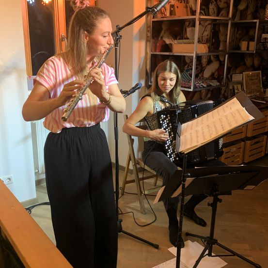 Zwei Frauen spielen in einem Raum, eine Flöte und die andere Akkordeon. Sie stehen vor einem Mikrofon und einem Notenständer.