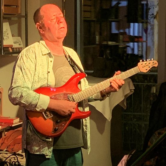 Ein Mann spielt eine rote E-Gitarre in einem Raum. Er trägt ein Blumenhemd und grüne Hosen.