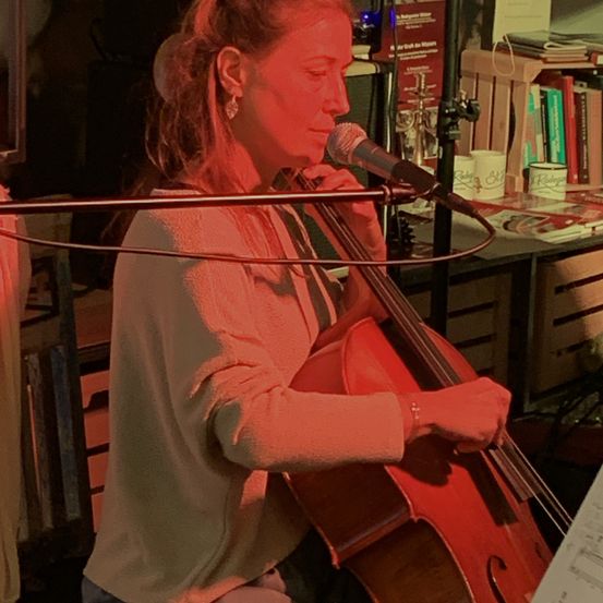 Eine Frau spielt Cello und singt in ein Mikrofon. Sie ist lässig gekleidet und hat einen ernsten Gesichtsausdruck. Das Cello ist an einem Mikrofonstativ angeschlossen. Hinter ihr steht ein Schreibtisch mit Büchern und anderen Gegenständen.