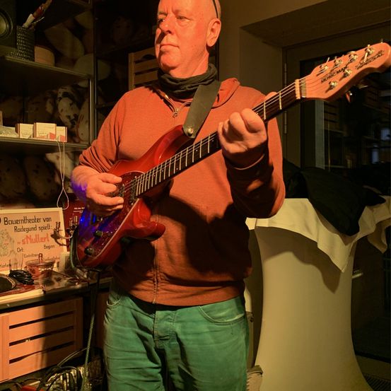 Ein Mann in einem braunen Shirt und grünen Hosen spielt eine rote E-Gitarre in einem schwach beleuchteten Raum mit Regalen und Kisten im Hintergrund.