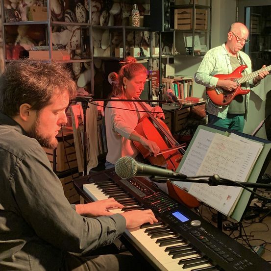 Drei Musiker sind in einem Raum, einer spielt Klavier, ein anderer Gitarre und eine Frau Cello. Sie üben wahrscheinlich für eine Aufführung.