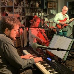 Drei Musiker sind in einem Raum, einer spielt Klavier, ein anderer Gitarre und eine Frau Cello. Sie üben wahrscheinlich für eine Aufführung.